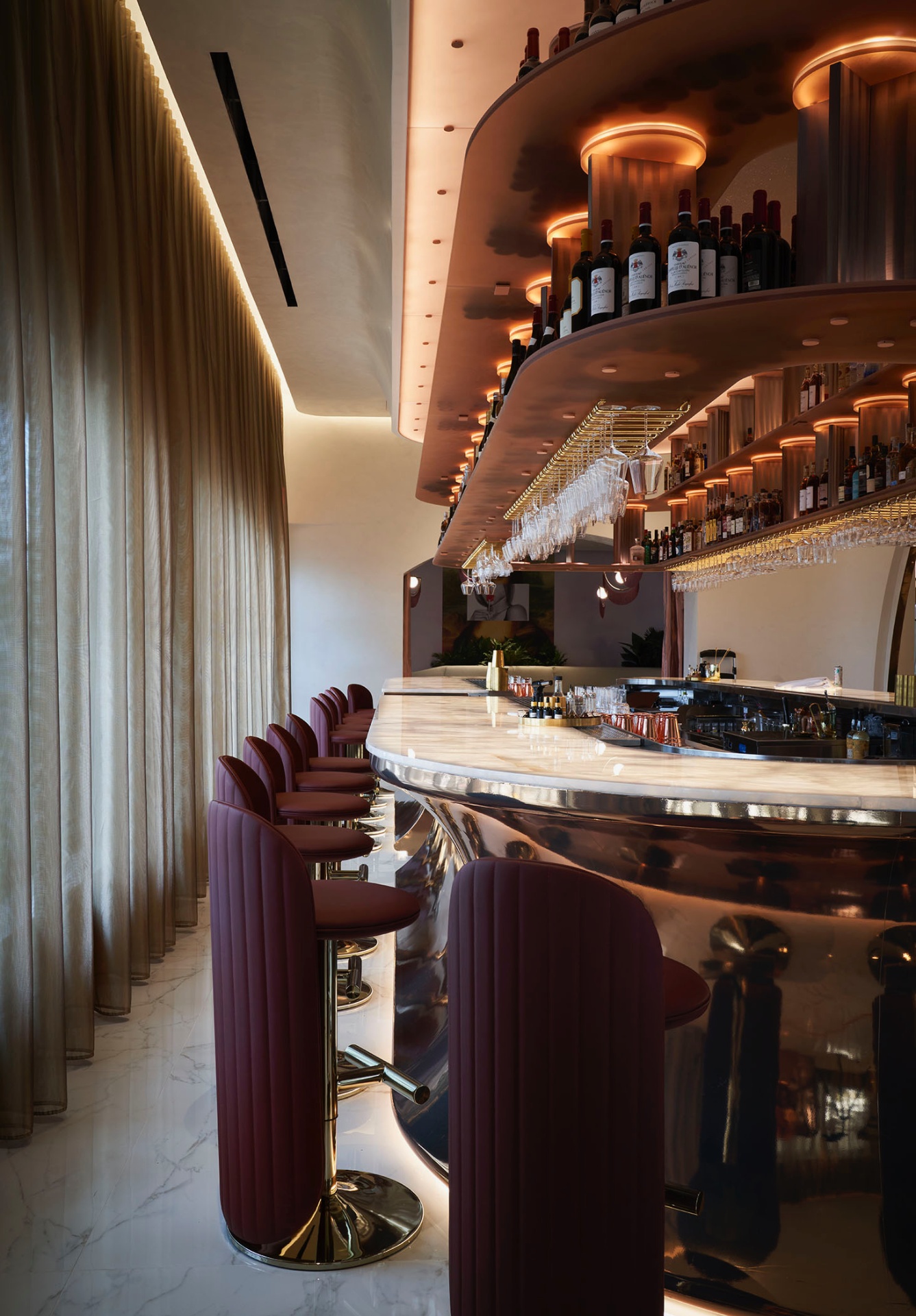 Curved marble bar at Cocody — burnished metal base, backlit tiered shelving of bottles and stemware, and a row of burgundy velvet stools set against sheer drapery.